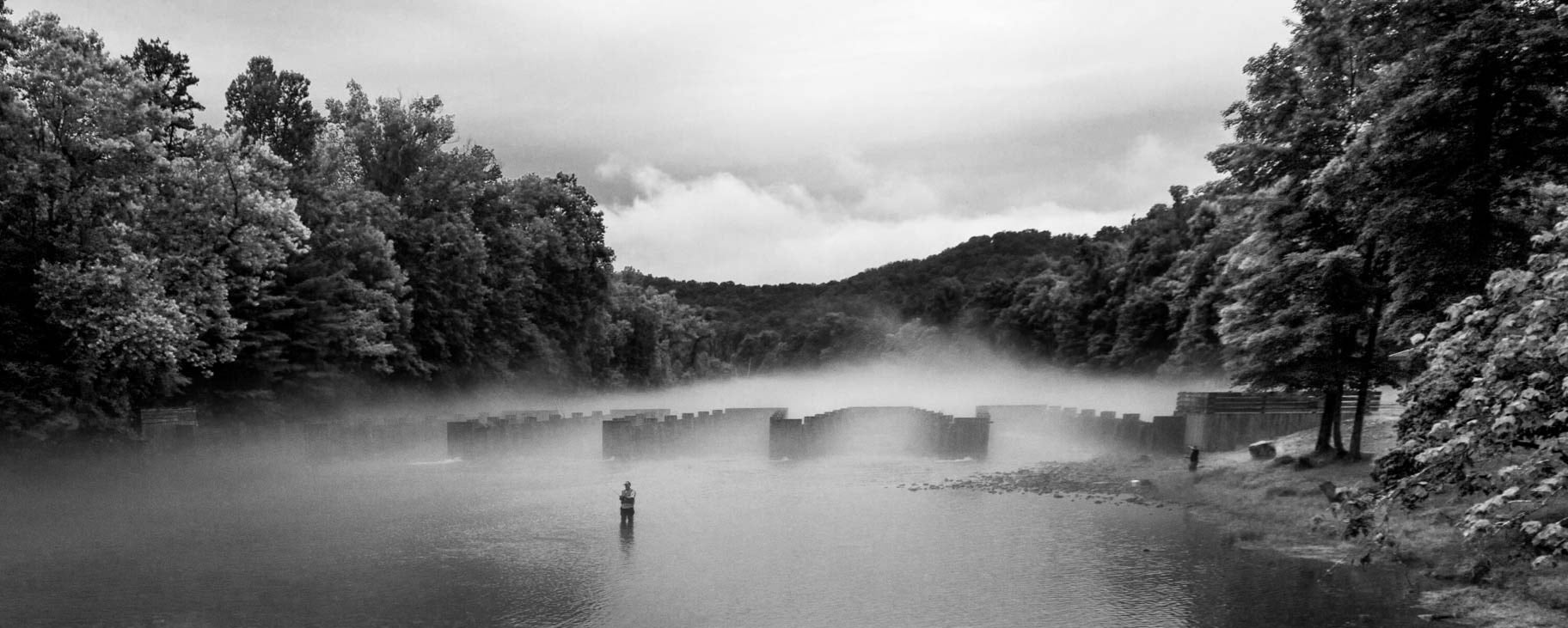 Black and white scene of a lake with a dam, trees, and a person fishing.