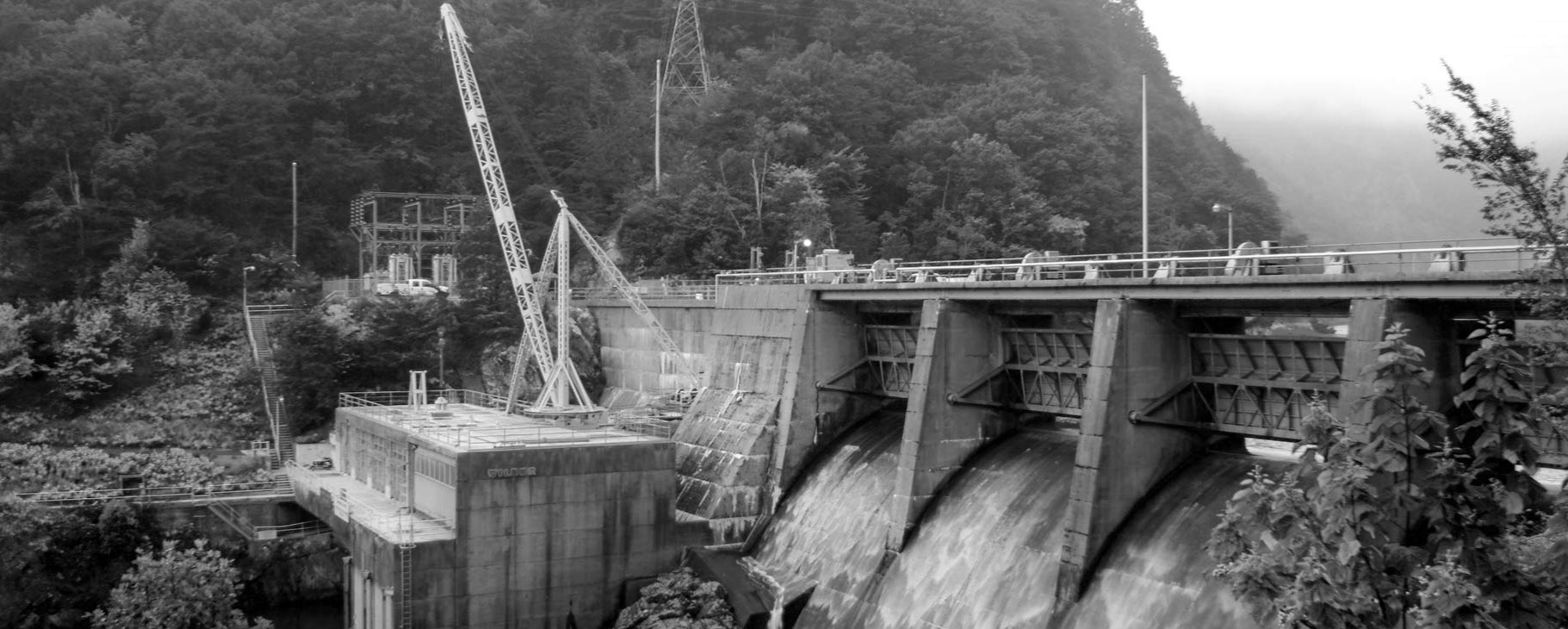 Black and white image of a dam with a crane and mountains in the background